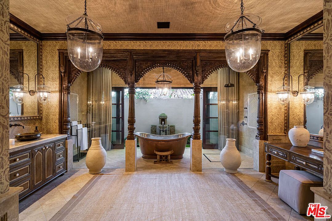 This luxurious primary bathroom features an ornate, carved wooden archway framing a deep copper soaking tub, creating a spa-like sanctuary. The room is adorned with textured wallpaper, elegant glass pendant lighting, and dual vanities with intricate cabinetry, all set against a backdrop of stone flooring and a private outdoor fountain view. The overall aesthetic is one of opulent, Mediterranean-inspired sophistication.
