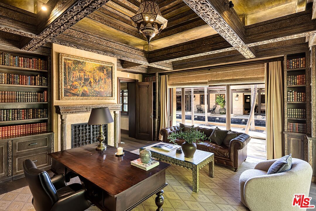 This sophisticated home office exudes a classic, scholarly atmosphere with its rich wood-paneled ceiling, extensive built-in bookshelves, and a grand fireplace. The room is furnished with a dark wood desk, a leather chesterfield sofa, and a plush armchair, all arranged to overlook a serene outdoor courtyard through large glass doors. The warm lighting and intricate architectural details create a luxurious and tranquil workspace.