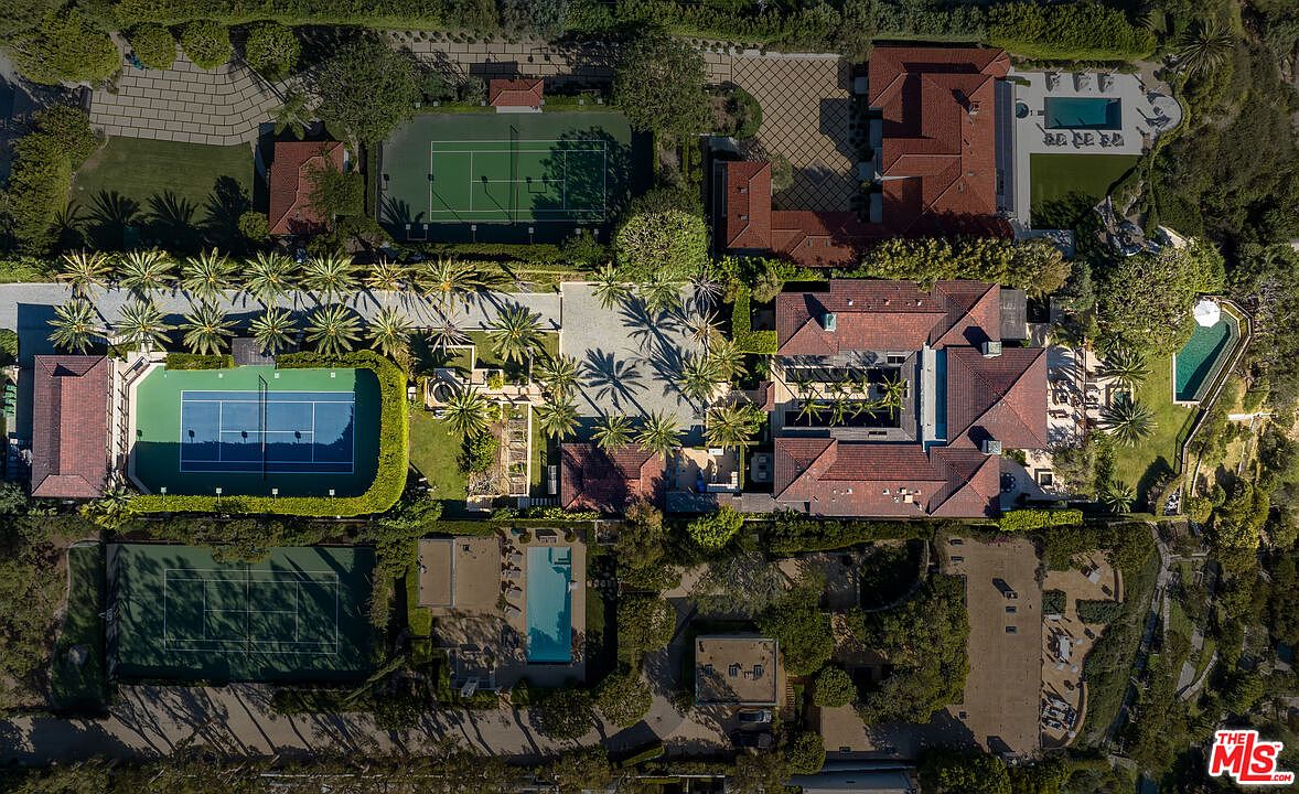 This expansive aerial view captures a luxurious estate featuring multiple tennis courts, swimming pools, and sprawling, meticulously landscaped grounds. The property showcases large, interconnected Mediterranean-style buildings with terracotta tile roofs, surrounded by lush greenery and mature palm trees. The perspective provides a comprehensive look at the vast scale and high-end amenities of this private compound.