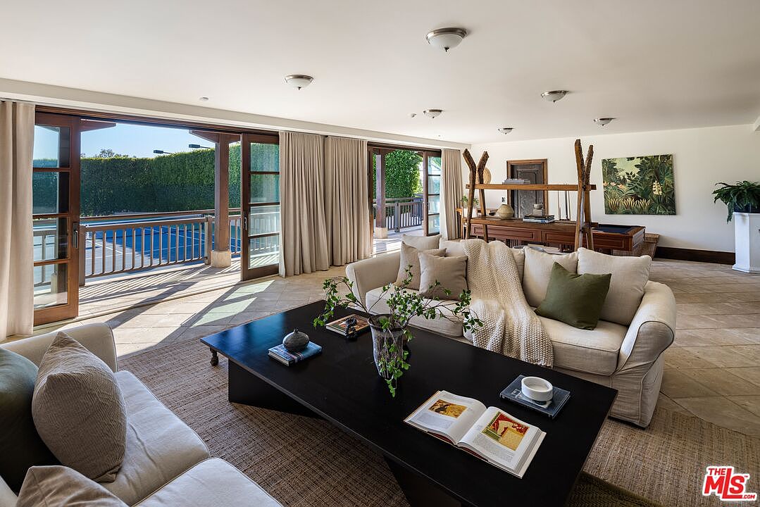 This spacious and airy living room features a neutral color palette with comfortable beige sofas and a large, dark wood coffee table as the centerpiece. Large sliding glass doors open onto a sun-drenched deck, seamlessly blending indoor and outdoor living spaces while offering a view of the pool area. The room is accented by natural textures, a unique wooden display structure, and tasteful decor, creating a serene and inviting atmosphere.