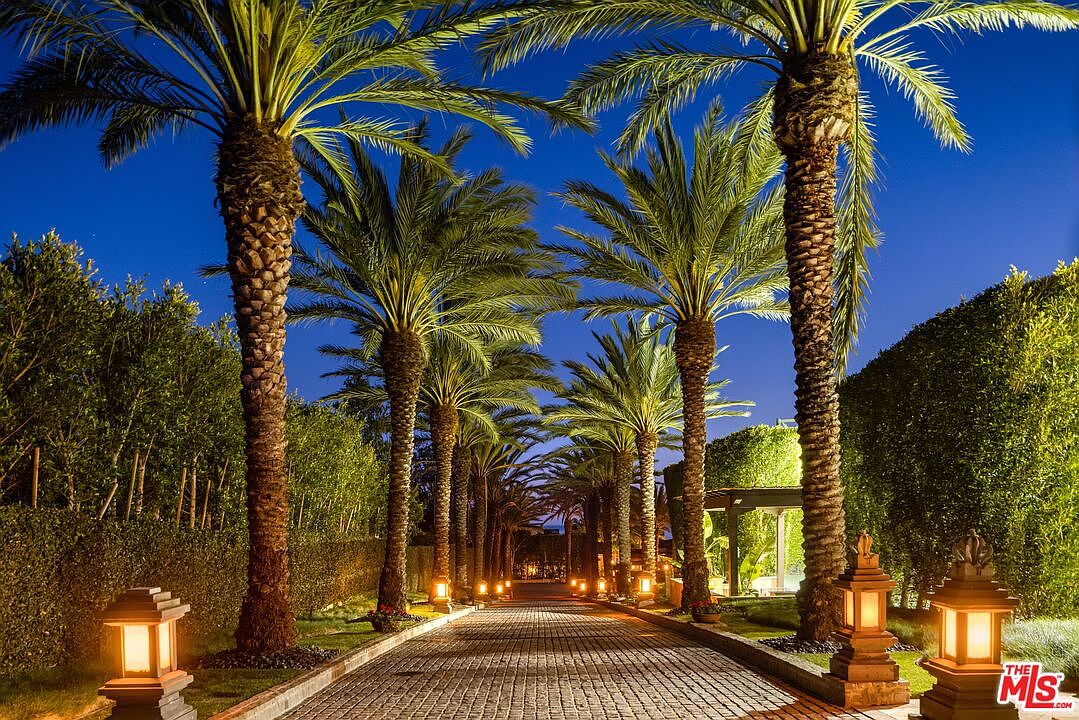 This cinematic shot captures a grand, palm-lined driveway at twilight, leading toward a private estate. The cobblestone path is illuminated by warm, traditional-style lanterns, creating a welcoming and luxurious atmosphere against the deep blue evening sky. The lush, manicured hedges and towering palm trees frame the perspective, emphasizing the property's exclusivity and sophisticated curb appeal.