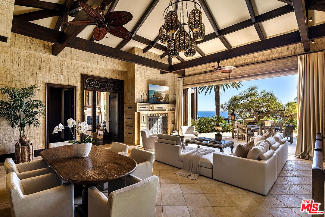 This expansive, open-concept living and dining area features a high vaulted ceiling with exposed dark wood beams and a striking multi-tiered chandelier. The space seamlessly transitions to an outdoor patio through large sliding glass doors, offering a stunning view of the ocean and lush greenery. The interior design blends tropical elegance with modern comfort, highlighted by a stone fireplace, neutral-toned plush furniture, and a rich wooden dining table.