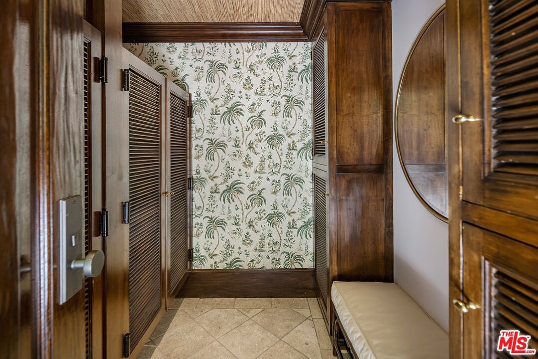 This interior hallway features a distinct tropical aesthetic with palm-patterned wallpaper and rich, dark wood louvered doors. A built-in bench with a neutral cushion sits beneath a circular mirror, while the floor is finished in light-toned stone tiles. The perspective is a straight-on view down the corridor, highlighting the contrast between the warm wood tones and the patterned wall covering.