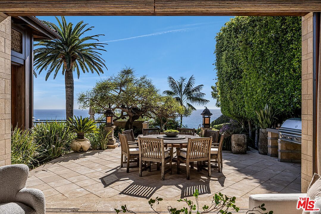 This luxurious outdoor patio offers a stunning, unobstructed view of the ocean, framed by lush greenery and a tall, manicured hedge. The space features a central wooden dining set surrounded by comfortable chairs, complemented by a built-in outdoor grill station to the right. The perspective is captured from a covered indoor-outdoor transition area, emphasizing the seamless blend between the home's interior and the serene coastal landscape.