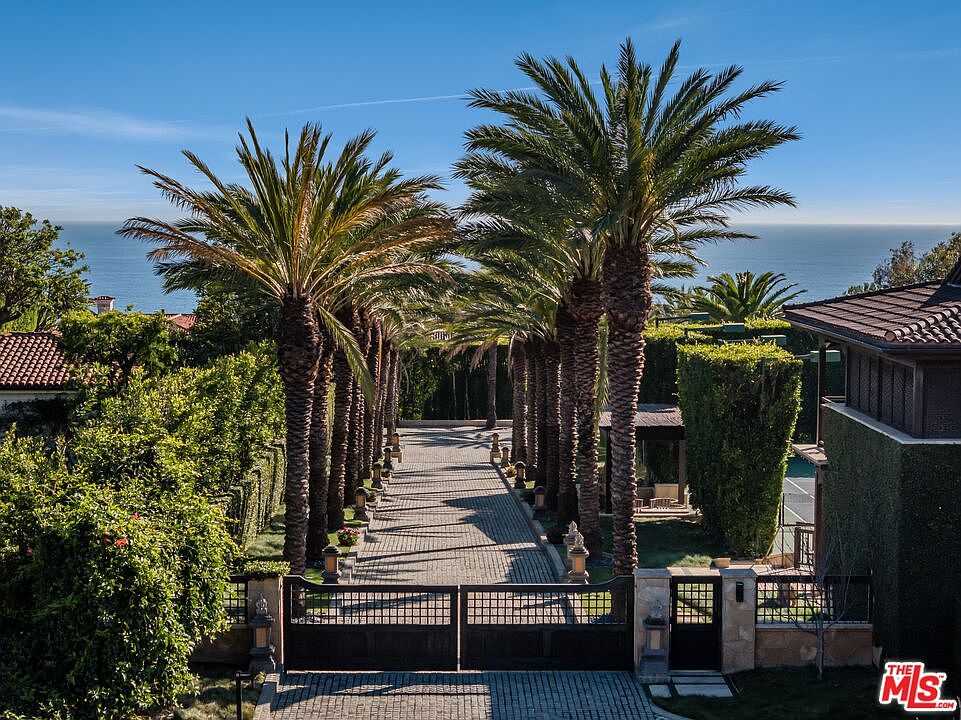 This grand gated entryway features a long, cobblestone driveway lined with mature, towering palm trees that create a dramatic, symmetrical corridor leading toward the ocean horizon. The scene is framed by lush, manicured hedges and stone pillars, evoking a sense of exclusive, coastal luxury. The perspective is elevated, capturing the impressive scale of the property's entrance and its stunning integration with the natural landscape.