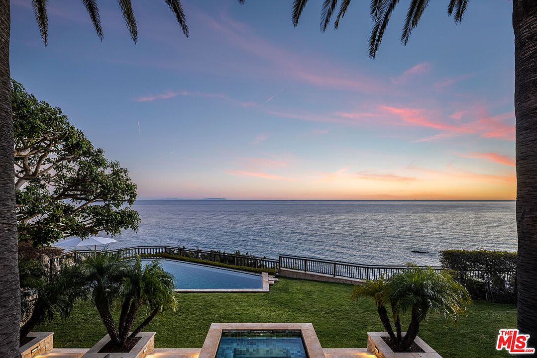 This stunning oceanfront property features a luxurious infinity-edge pool and a foreground spa, seamlessly blending with the vast horizon of the Pacific Ocean. The scene is captured at sunset, with a vibrant, soft-hued sky casting a serene glow over the manicured lawn and lush palm trees. The elevated perspective emphasizes the dramatic coastal location and the tranquil, high-end lifestyle offered by the residence.