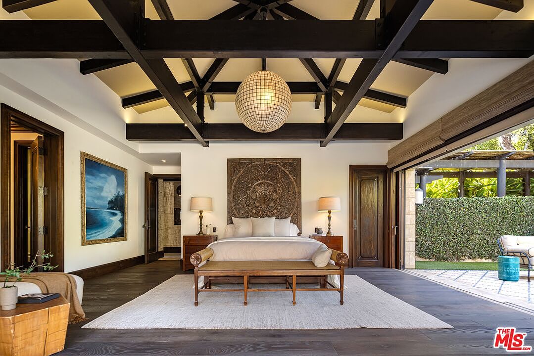 This luxurious primary bedroom features a dramatic vaulted ceiling with exposed dark wood beams and a large, spherical pendant light. The room is anchored by a king-sized bed with a striking carved wooden headboard, flanked by matching nightstands and lamps, and opens directly onto a private patio through expansive sliding glass doors. The aesthetic blends tropical elegance with high-end design, creating a serene and sophisticated retreat.