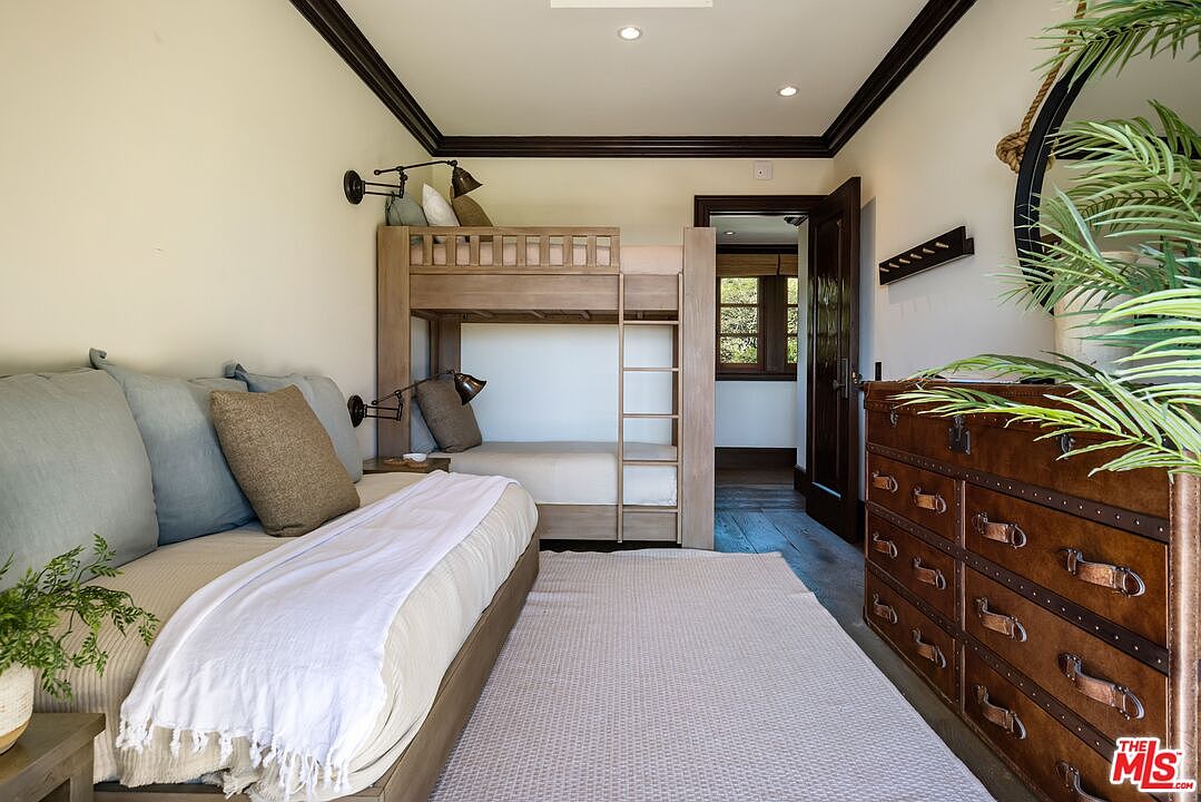 This cozy bedroom features a rustic, wood-framed bunk bed and a daybed with soft blue and neutral-toned bedding, creating a warm and inviting atmosphere. The room is accented by a dark wood dresser with leather-strap pulls and a textured area rug, all set against light-colored walls with dark crown molding. The perspective captures a comfortable, multi-functional sleeping space that feels both stylish and practical for guests or children.