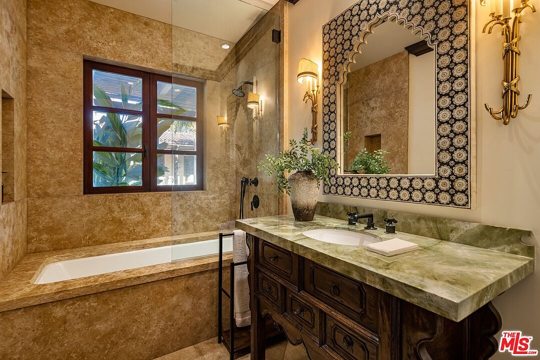 This luxurious primary bathroom features a rich, Mediterranean-inspired aesthetic with warm travertine stone walls and a striking green onyx vanity countertop. The space includes a deep soaking tub integrated into the stone surround, a decorative arched mirror with intricate tile detailing, and ornate gold-toned wall sconces. The perspective captures the elegant blend of rustic textures and sophisticated finishes, creating a spa-like atmosphere.