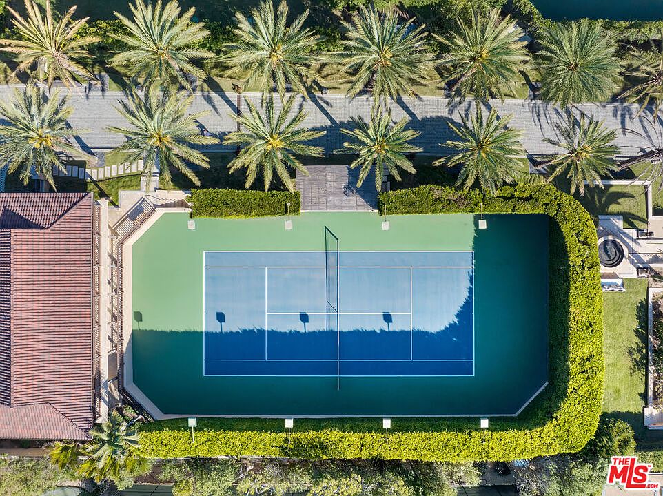 This high-angle aerial view captures a private tennis court surrounded by lush, manicured hedges and a row of tall palm trees. The court features a vibrant blue playing surface with green borders, set against a backdrop of a paved walkway and a portion of a residential roof. The perspective emphasizes the property's expansive outdoor recreational space and well-maintained landscaping.