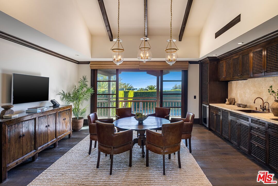 This elegant dining room features a warm, rustic aesthetic with dark wood cabinetry, exposed ceiling beams, and a round wooden table surrounded by leather-upholstered chairs. A large sliding glass door provides a seamless transition to an outdoor balcony, bathing the room in natural light and offering a scenic view. The space is anchored by a textured area rug and three sophisticated glass pendant lights, creating a cozy yet refined atmosphere.