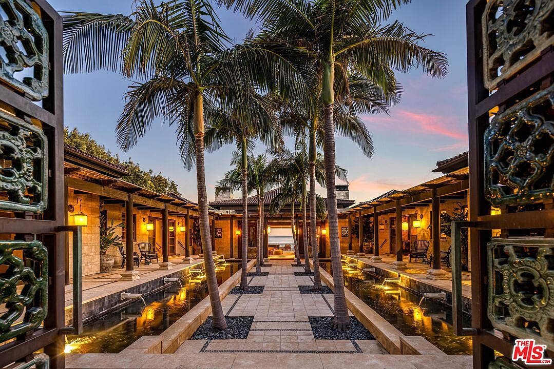 This stunning entryway features a symmetrical courtyard design with a central stone walkway flanked by two long, illuminated reflecting pools. Tall, mature palm trees rise from the water features, leading the eye toward a framed view of the ocean in the distance. The architectural style is sophisticated and resort-like, framed by ornate, dark metal gates that create a dramatic, cinematic perspective.