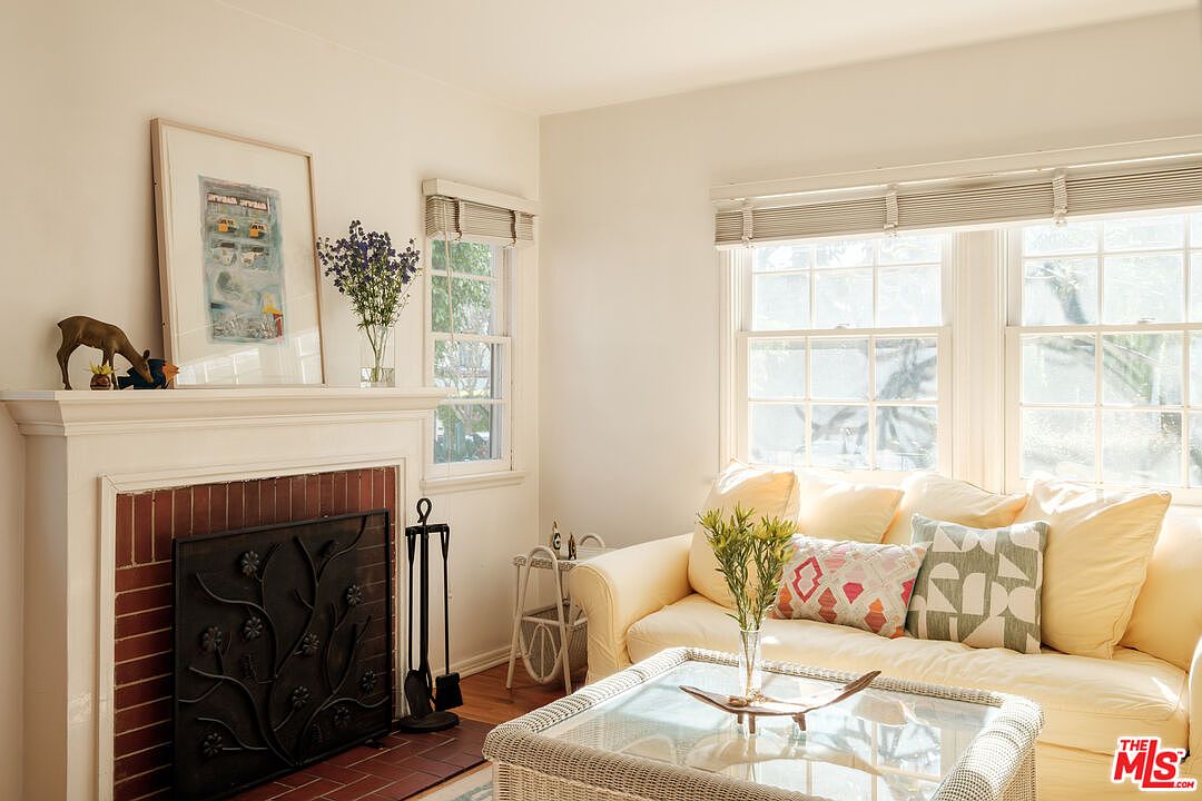 This is a cozy living room featuring a fireplace with a decorative screen and mantel, a comfortable yellow sofa with patterned pillows, and a glass-topped wicker coffee table. Natural light streams in through the windows, creating a warm and inviting atmosphere. The room is decorated in a traditional style with neutral walls and wood floors.