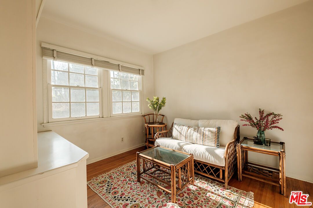 This cozy living room features a rattan sofa with white cushions and a patterned throw pillow, complemented by matching rattan side tables and a coffee table with glass tops. A colorful area rug adds warmth to the hardwood floors, while natural light streams in through the windows, creating a bright and inviting atmosphere. The room is decorated with floral arrangements, enhancing the homey feel.