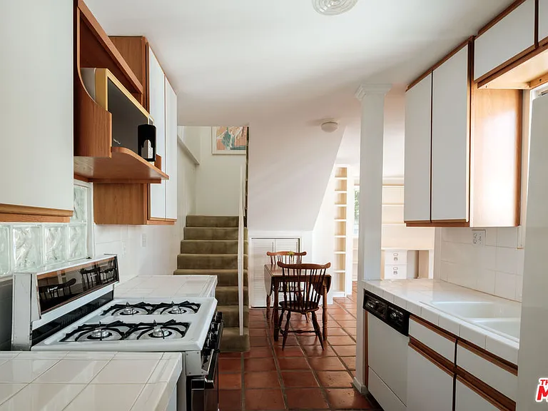 This is an interior shot of a kitchen area that leads into a dining space and staircase. The kitchen features white cabinets with wooden trim, white tiled countertops, and a white range. The floors are terracotta tile, adding warmth to the space.