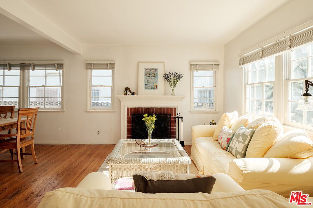This is a bright and inviting living room featuring hardwood floors, white walls, and large windows that let in plenty of natural light. The room is furnished with a comfortable yellow sofa adorned with decorative pillows, a wicker coffee table with a glass top, and a fireplace with a white mantel. The overall style is cozy and traditional, creating a warm and welcoming atmosphere.