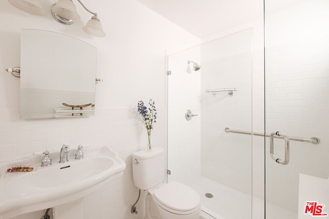 This is a bright, clean bathroom featuring white subway tile walls and a glass-enclosed shower. A classic pedestal sink with chrome fixtures and a toilet are visible, along with a vase of blue flowers adding a touch of color. The overall impression is fresh and well-maintained.