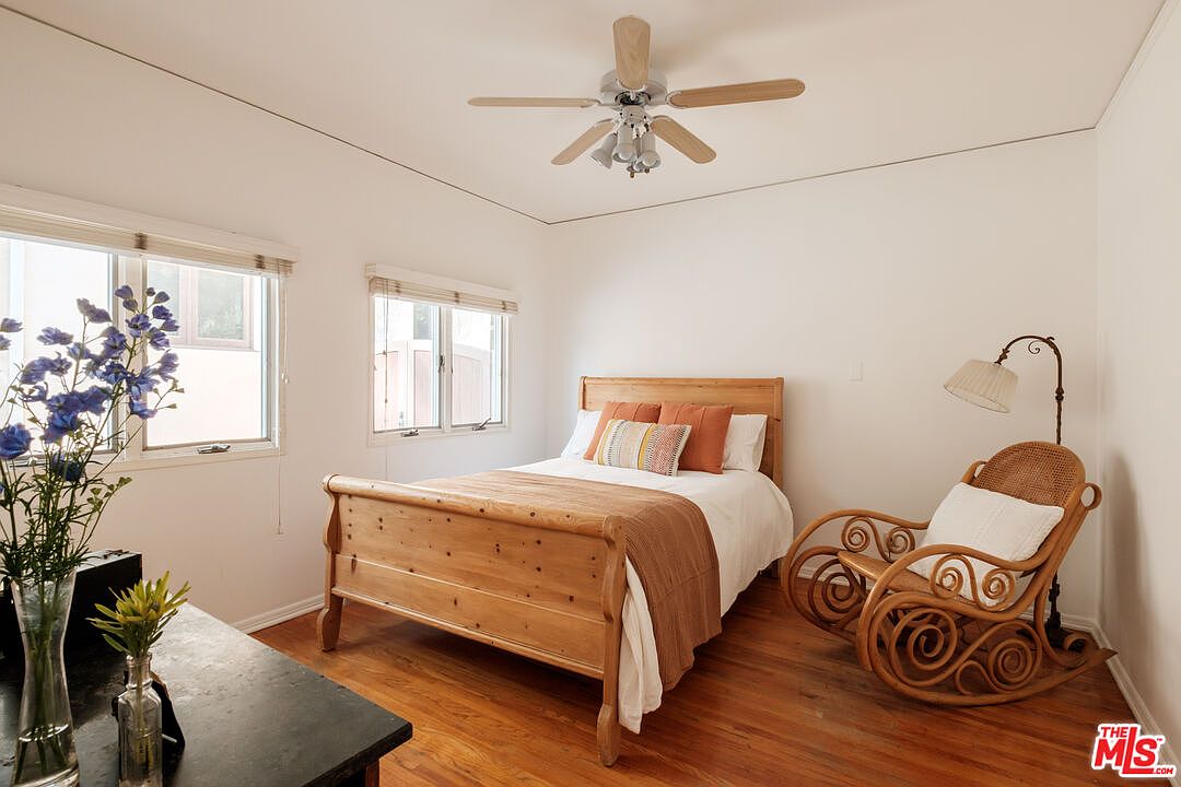This is a cozy bedroom featuring a wooden bed frame with orange and patterned pillows, a white comforter, and a brown throw blanket. A wicker rocking chair sits beside the bed, illuminated by a floor lamp. The room has hardwood floors and white walls, with natural light streaming in through two windows.