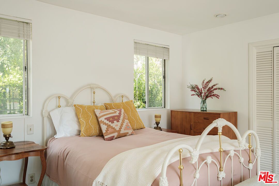 This is a bright and airy bedroom featuring a white iron bed with gold accents, adorned with a pink quilt, white throw blanket, and decorative pillows. Natural light streams in through two windows, complementing the white walls and creating a serene atmosphere. A wooden dresser and bedside table add warmth and character to the space.