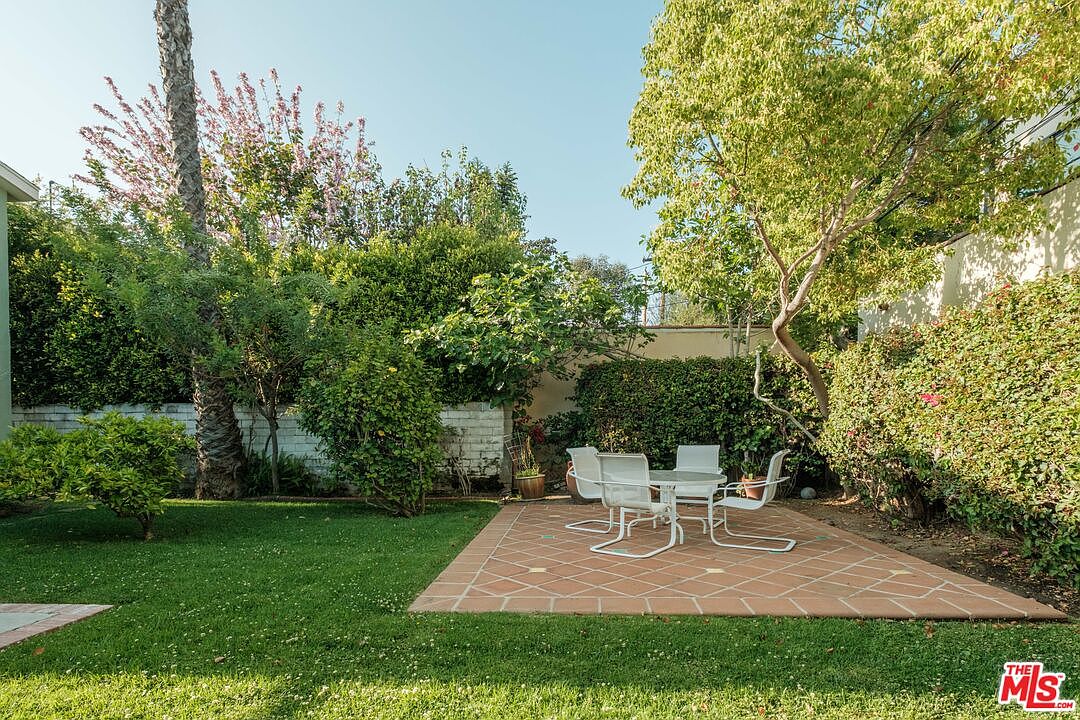 This image showcases a well-maintained backyard garden with lush green grass and various trees and bushes. A brick patio area features a white outdoor dining set, creating an inviting space for relaxation and entertainment. The yard is enclosed by a wall and mature hedges, providing privacy and a serene atmosphere.
