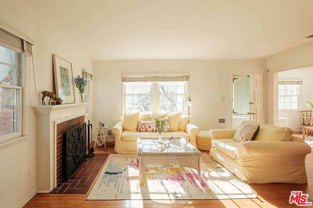 The living room features a cozy and inviting atmosphere with two light yellow sofas, a wicker coffee table with a glass top, and a fireplace with a decorative screen. Natural light floods the room through large windows, highlighting the hardwood floors and a colorful area rug. The overall impression is one of comfort and classic elegance.