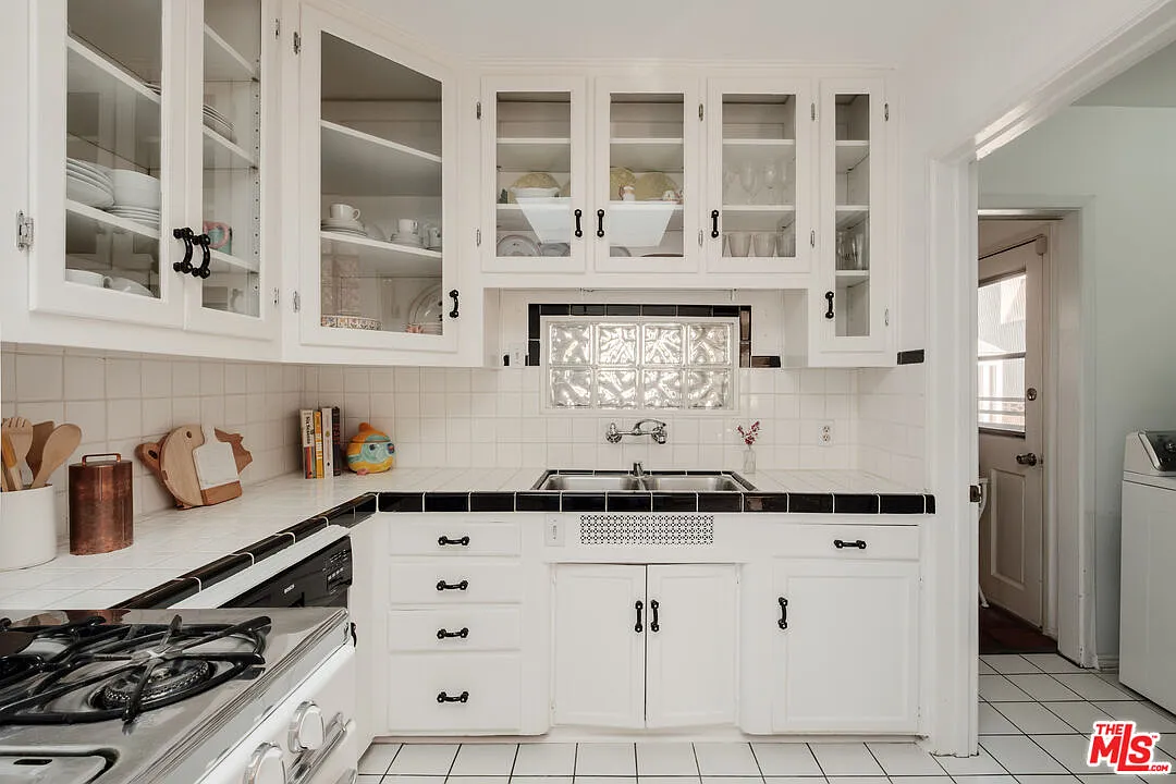 This kitchen showcases classic style with white cabinetry, black countertops, and tiled backsplash and flooring. Glass-front cabinets display dishware, while lower cabinets and drawers provide ample storage. The kitchen is well-lit and appears clean, providing a functional space for cooking and meal preparation.
