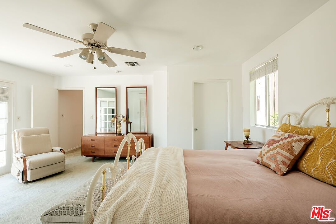 This is a bright and airy primary bedroom featuring white walls, a vintage-style metal bed frame with a light pink bedspread, and a ceiling fan. A wooden dresser with two mirrors sits against the wall, and a comfortable armchair is positioned near a door leading to the outside. The room has a calm and inviting atmosphere.