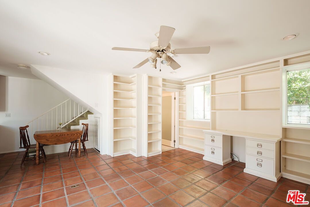 This interior shot showcases a well-lit office or study area with terracotta tile flooring. The room features built-in bookshelves and a desk area with drawers, providing ample storage and workspace. A ceiling fan adds to the room's functionality, while the staircase in the background suggests a multi-level home.