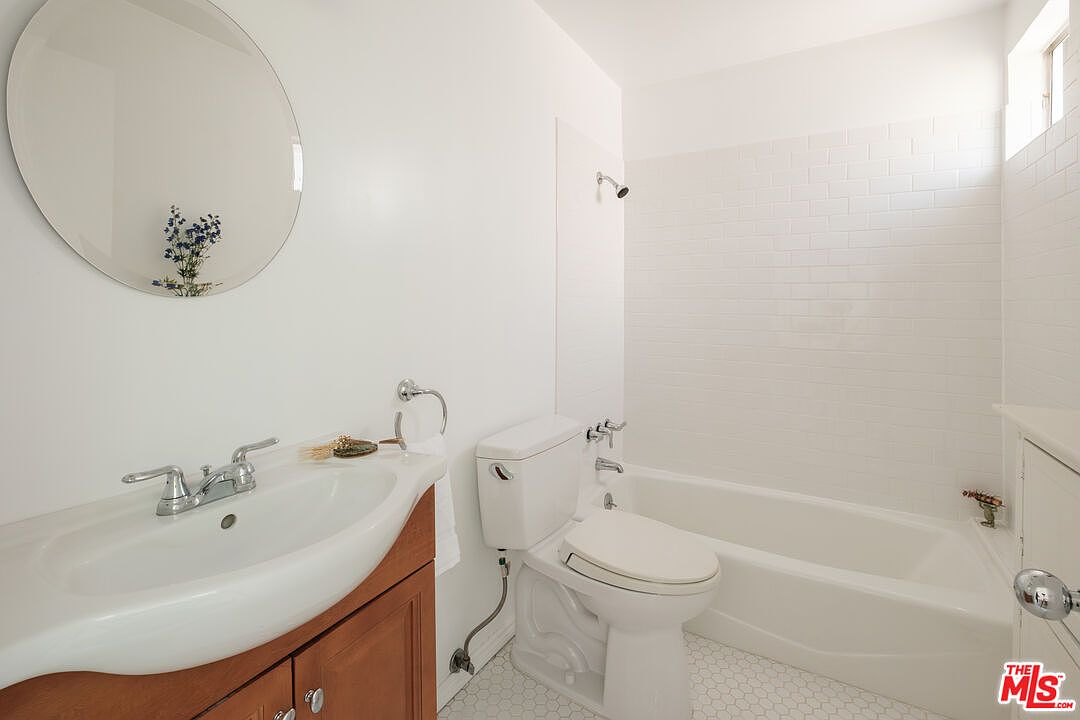 This is a bright and clean bathroom featuring a white toilet, a white sink with a wood-toned vanity, and a round mirror above the sink. The bathtub and shower area are tiled in white, creating a fresh and modern look. The floor is covered in small, white hexagonal tiles.