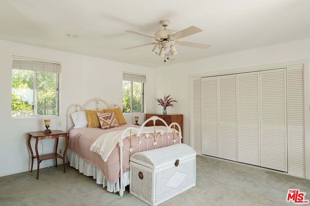 This is a bright and airy bedroom featuring a white iron bed frame with pink bedding and decorative pillows. A white wicker chest sits at the foot of the bed, and a small wooden side table is positioned next to the bed. The room has white walls, light-colored carpet, and a large closet with louvered doors, creating a clean and inviting space.