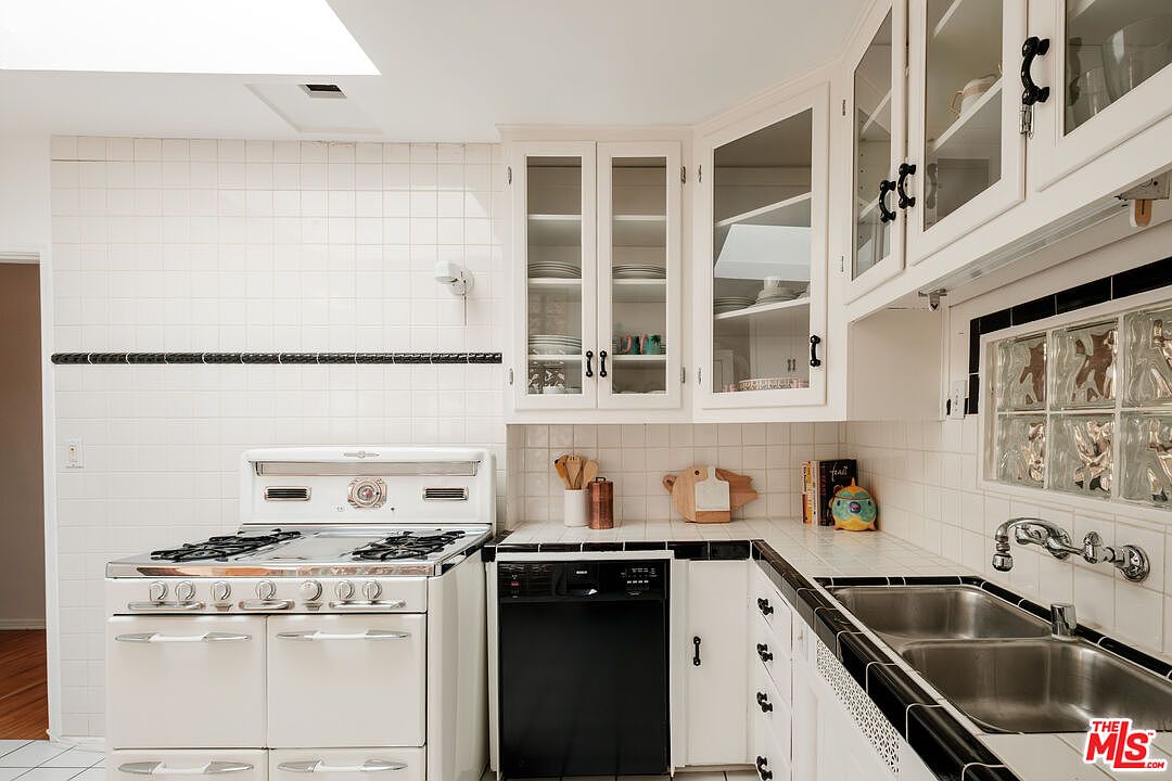 This is a well-lit kitchen featuring white tiled walls and countertops with black trim. The kitchen includes a vintage white stove, a black dishwasher, and white cabinets, some with glass doors. A stainless steel sink is visible, and the overall style is retro with a clean and functional design.