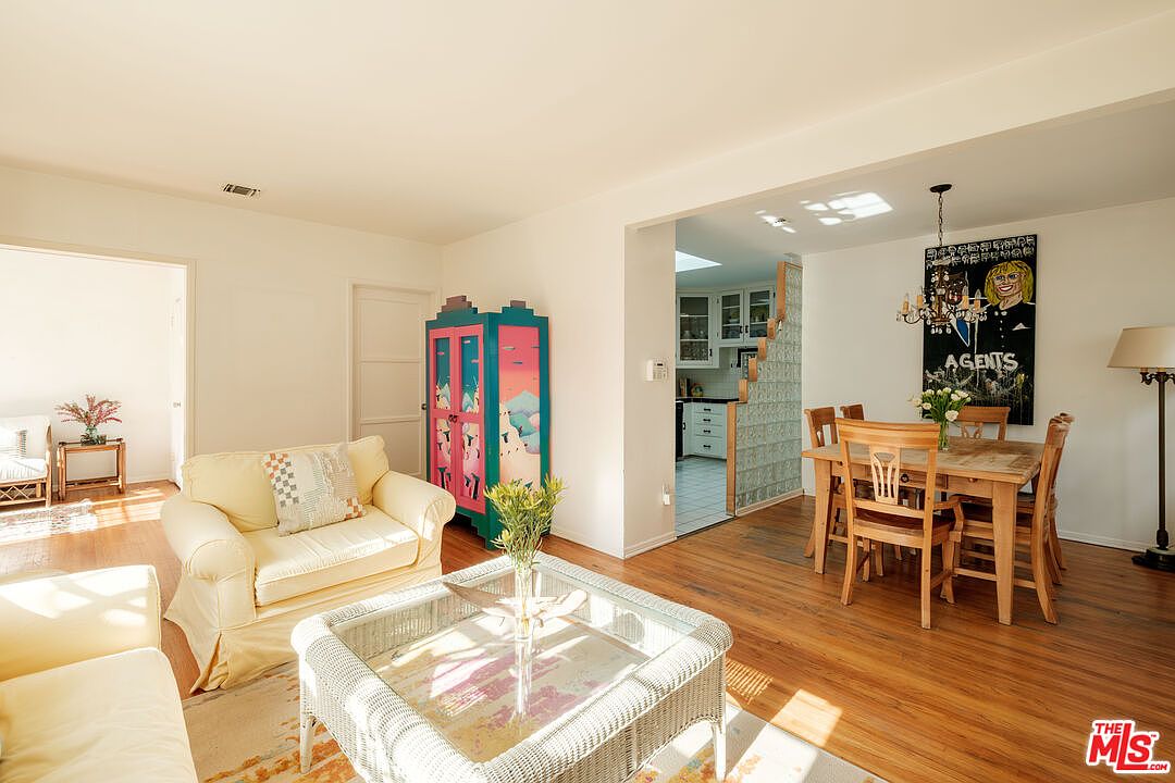 This is an interior shot of a living room and dining area. The living room features a comfortable yellow armchair, a wicker coffee table, and a colorful cabinet. The dining area includes a wooden table with chairs and a unique piece of art on the wall. The hardwood floors and natural light create a warm and inviting atmosphere.