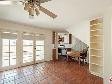 The image showcases a living room area with terracotta tile flooring and white walls. French patio doors provide access to an exterior space and natural light. A dining table with chairs is visible in front of an open kitchen, and a built-in bookcase adds to the room's storage and character.