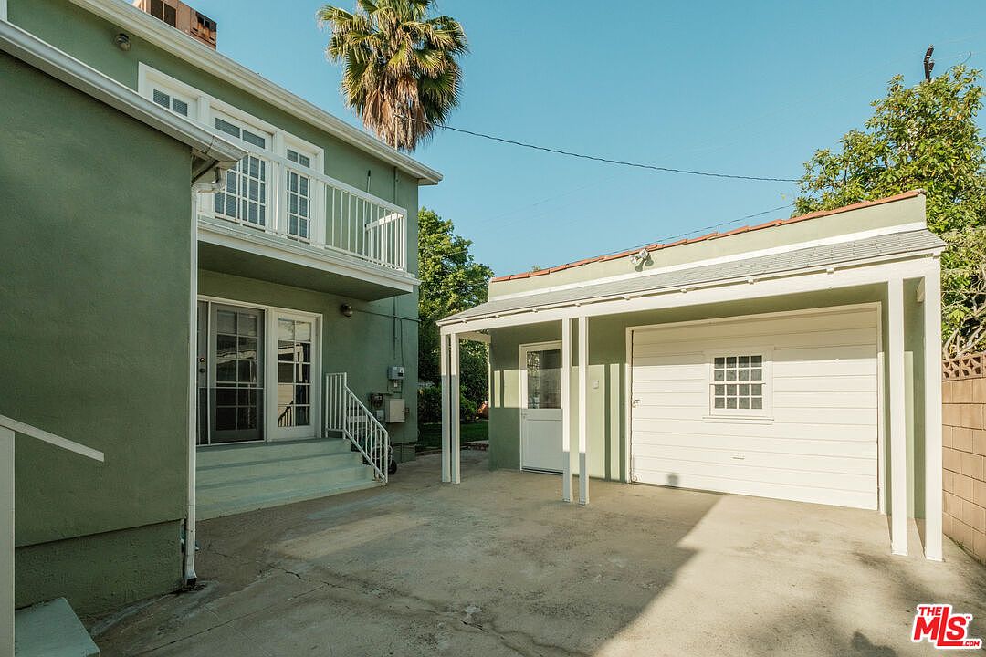 This exterior shot showcases the rear of a two-story house with a balcony and a detached garage. The house and garage are painted in a similar light green color, creating a cohesive look. The backyard features a concrete patio area, and the overall impression is a well-maintained and private outdoor space.