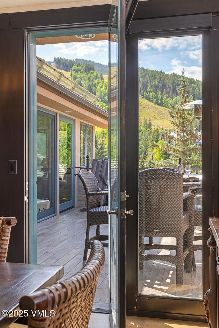 This image showcases a patio or balcony accessible through large, modern glass doors. The outdoor space features wicker chairs and a table set for dining, suggesting a comfortable area for relaxation and entertainment. The backdrop includes a scenic view of a tree-covered hillside, enhancing the property's appeal with its natural surroundings.