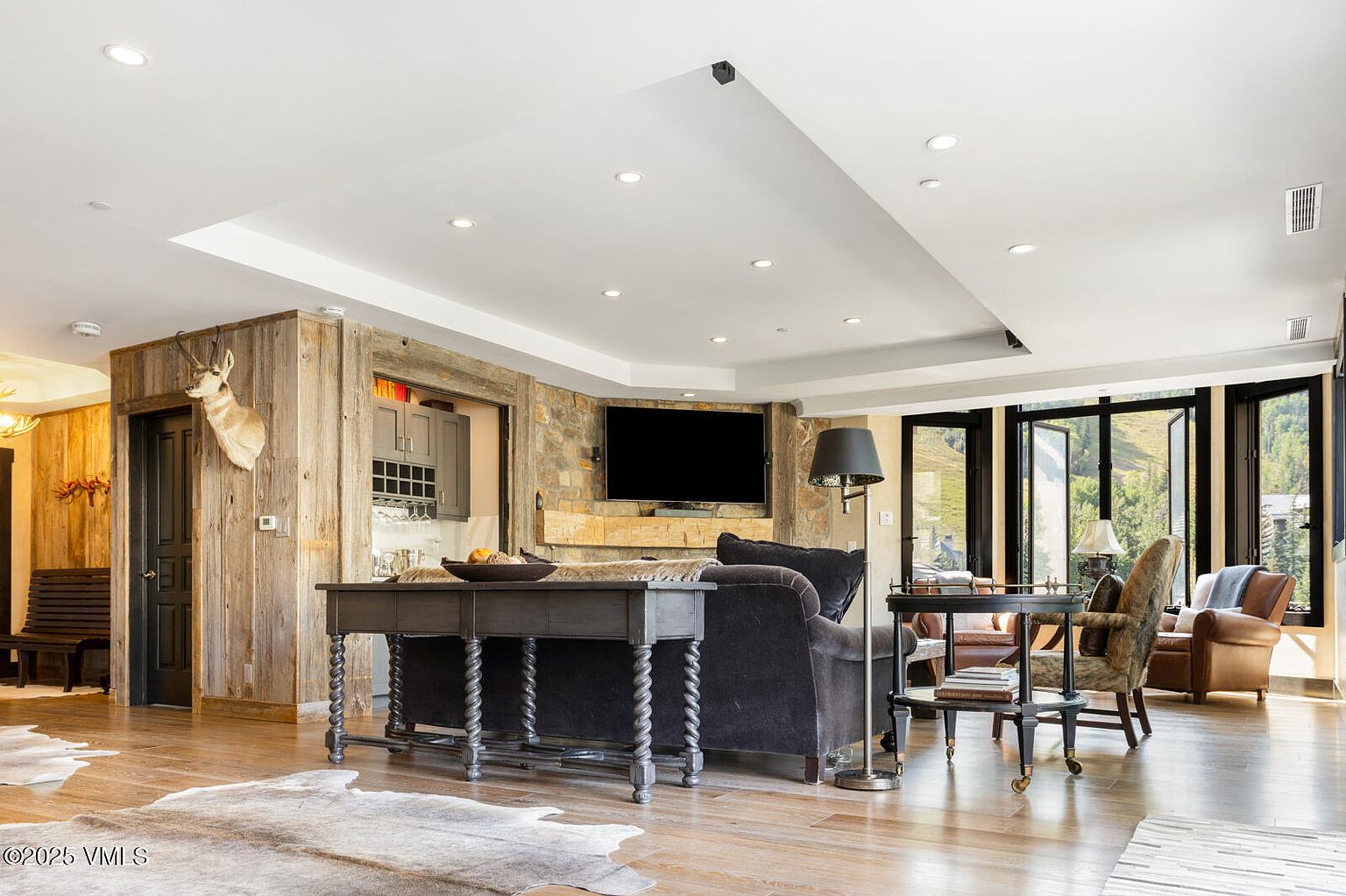 This is an interior shot of a living room featuring a blend of rustic and modern design elements. The room includes a stone fireplace with a mounted television, a dark-colored sofa, and a wooden console table. Large windows offer a view of the outdoors, and the flooring appears to be hardwood, complemented by a cowhide rug.