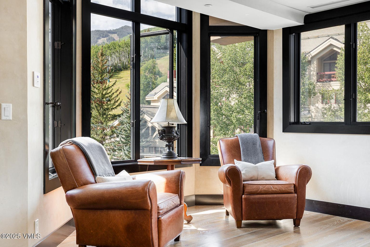 This sunroom features two leather armchairs positioned to enjoy the views from the large windows. The windows are framed in black and offer views of the surrounding landscape, including trees and a distant mountain. A small table with a lamp sits between the chairs, creating a cozy and inviting atmosphere.