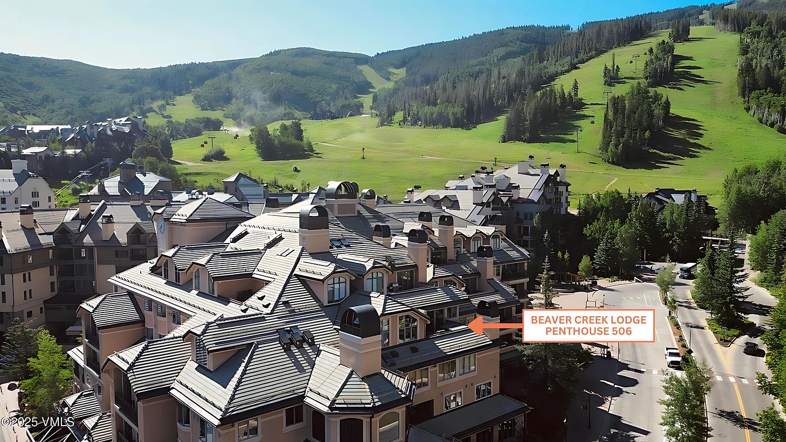 This aerial view showcases a luxurious residential complex nestled in a mountainous landscape. The architecture features steeply pitched roofs with multiple dormers and chimneys, suggesting high-end construction and design. The surrounding environment includes lush green slopes, indicating a desirable location for outdoor activities and scenic views.