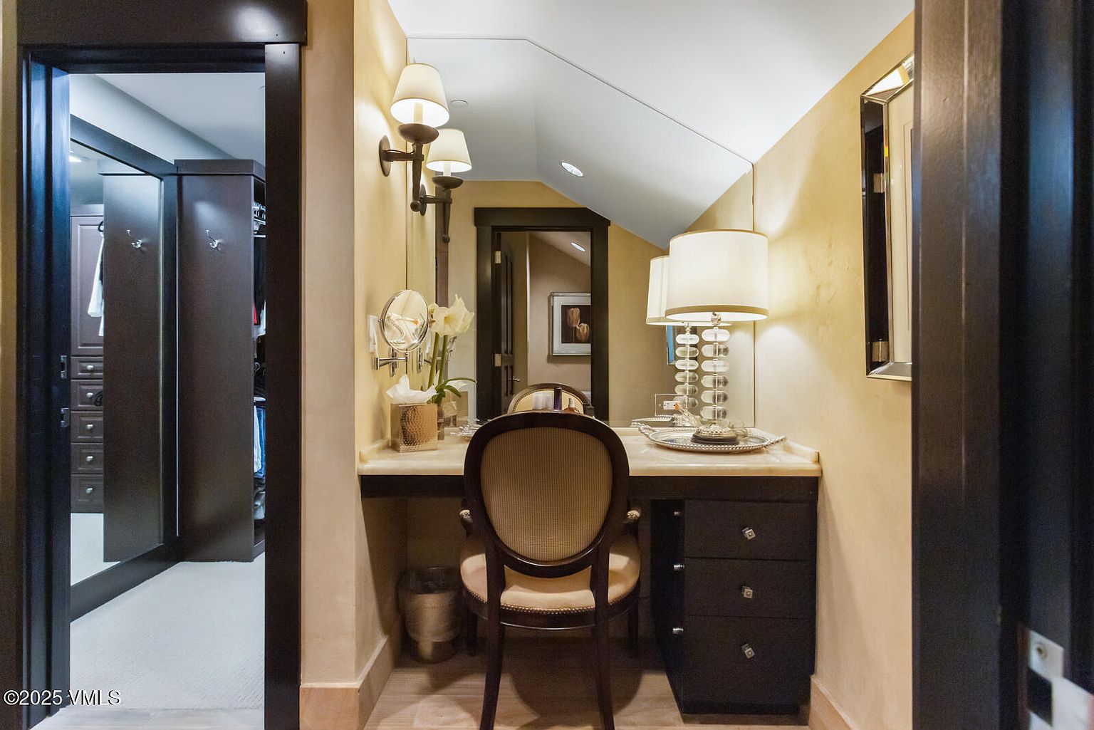 This is a well-appointed primary bathroom featuring a vanity area with a chair, a mirror, and decorative lighting. The color palette is warm and inviting, with beige walls and dark wood accents. The space appears luxurious and well-organized, suggesting a comfortable and functional area for personal care.