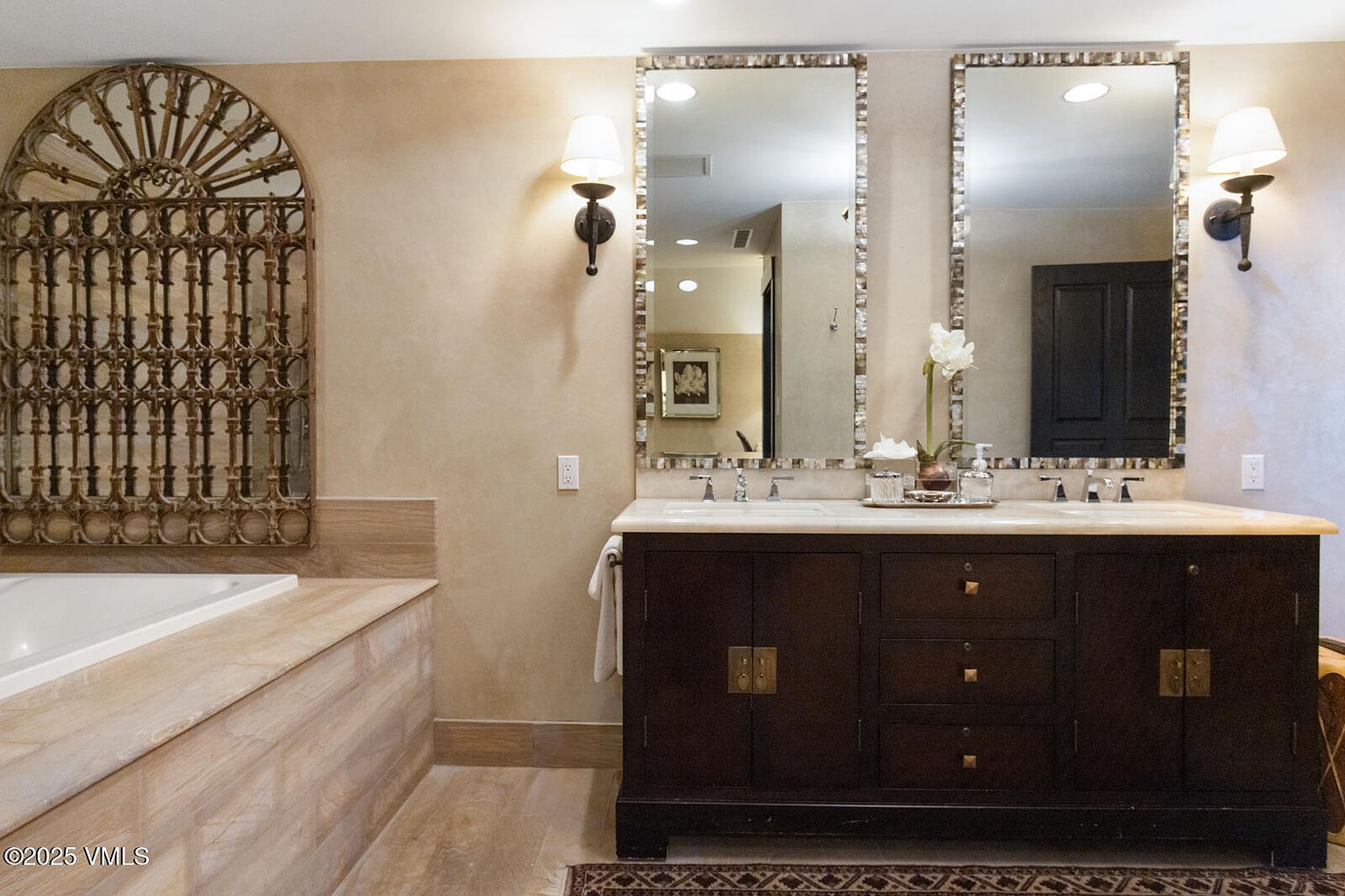 This is a primary bathroom featuring a double vanity with dark wood cabinetry and a light countertop. Two decorative mirrors with mosaic-style frames hang above the sinks, flanked by sconce lighting. A large, ornate metal piece adorns the wall above the bathtub, adding a unique architectural element to the space.