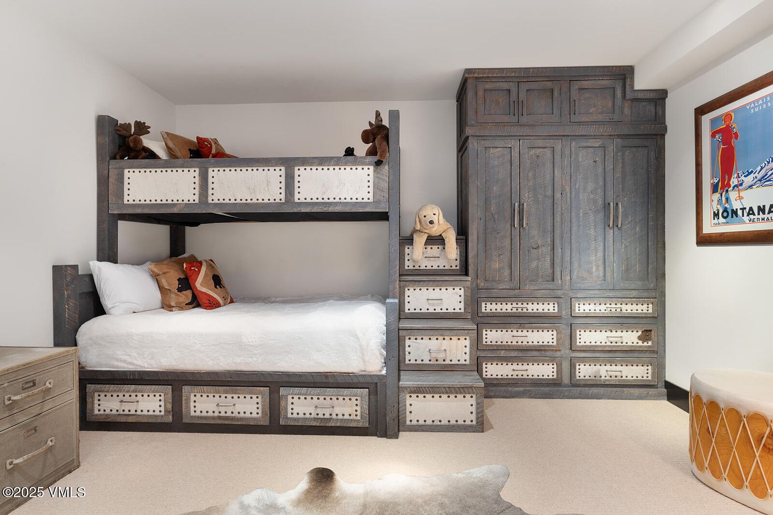 This is a cozy bedroom featuring a rustic bunk bed with built-in storage drawers and a matching wardrobe. The room has a warm and inviting feel, with neutral-toned walls and carpet. The bunk bed is adorned with decorative pillows and stuffed animals, adding a playful touch.