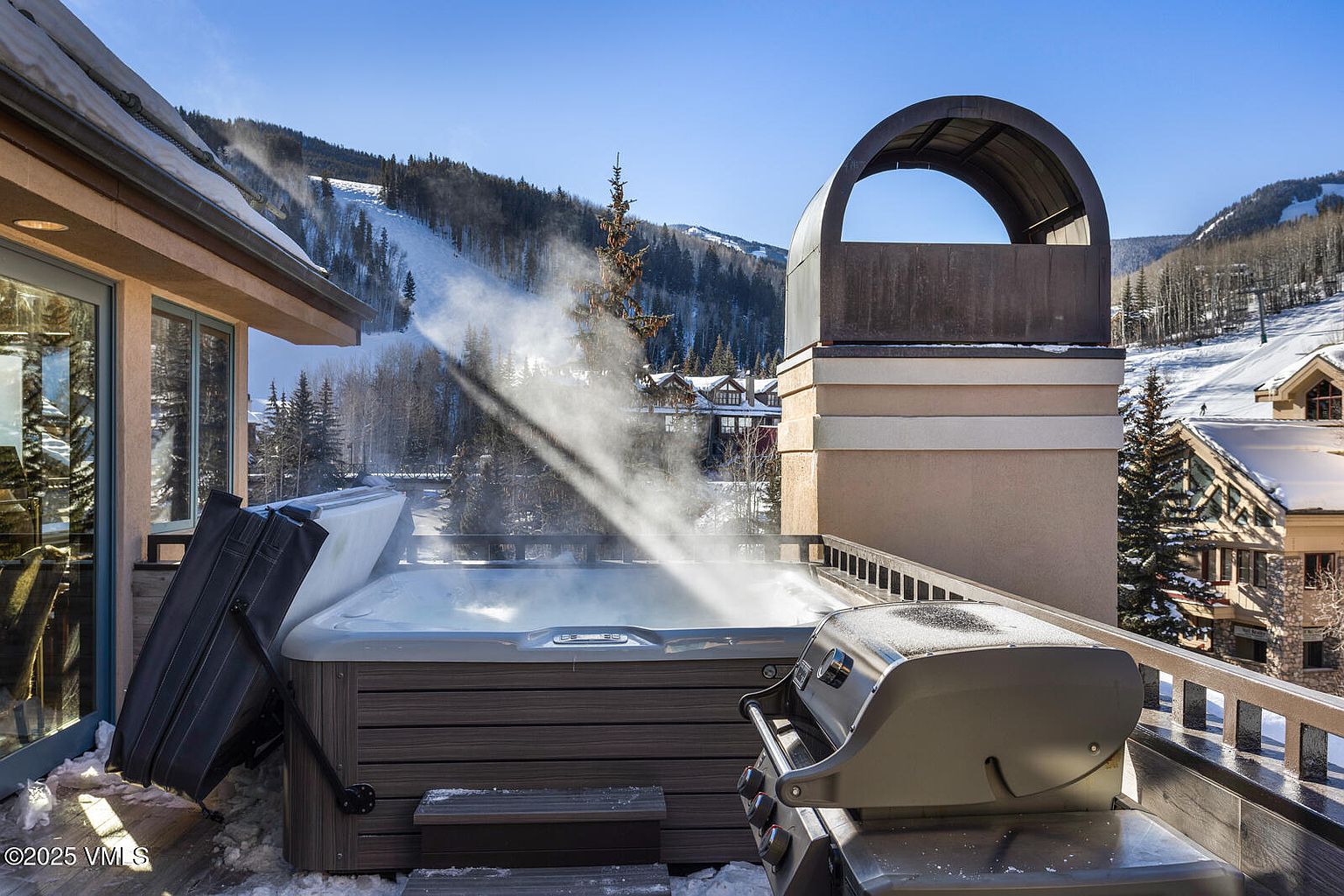 This image showcases a luxurious outdoor patio or deck area, complete with a hot tub and a grill, suggesting a space designed for relaxation and entertainment. The backdrop features a scenic mountain view, possibly a ski slope, enhancing the property's appeal for winter sports enthusiasts. The presence of snow and the architectural style of the surrounding buildings indicate a high-end resort or residential area.