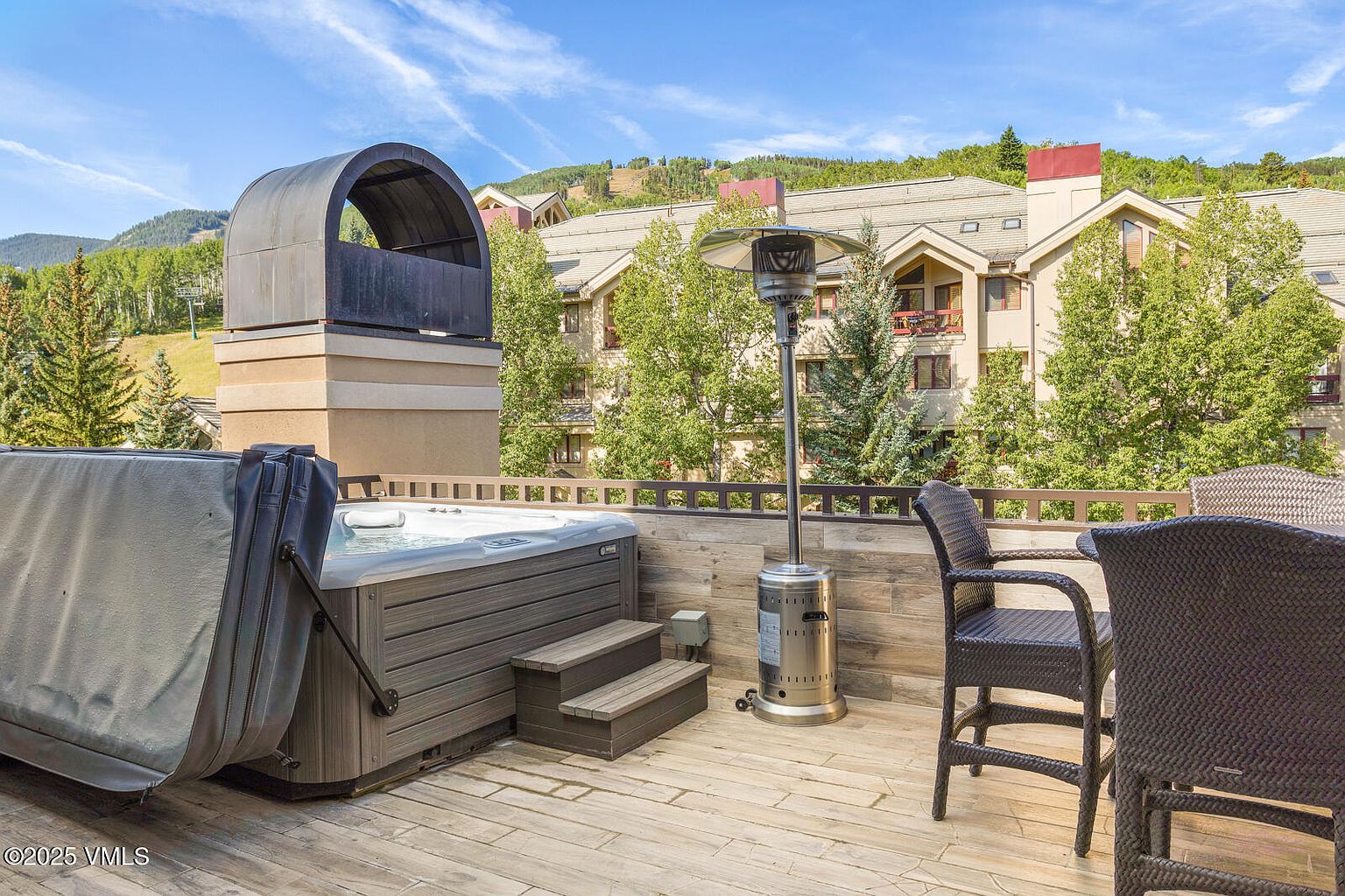 This image showcases a luxurious outdoor patio or deck area, featuring a hot tub with a cover partially open, a modern patio heater, and comfortable wicker chairs. The deck is constructed with light-colored wooden planks, and the backdrop includes a multi-story building with trees, suggesting a resort or upscale residential setting. The scene evokes a sense of relaxation and high-end outdoor living.
