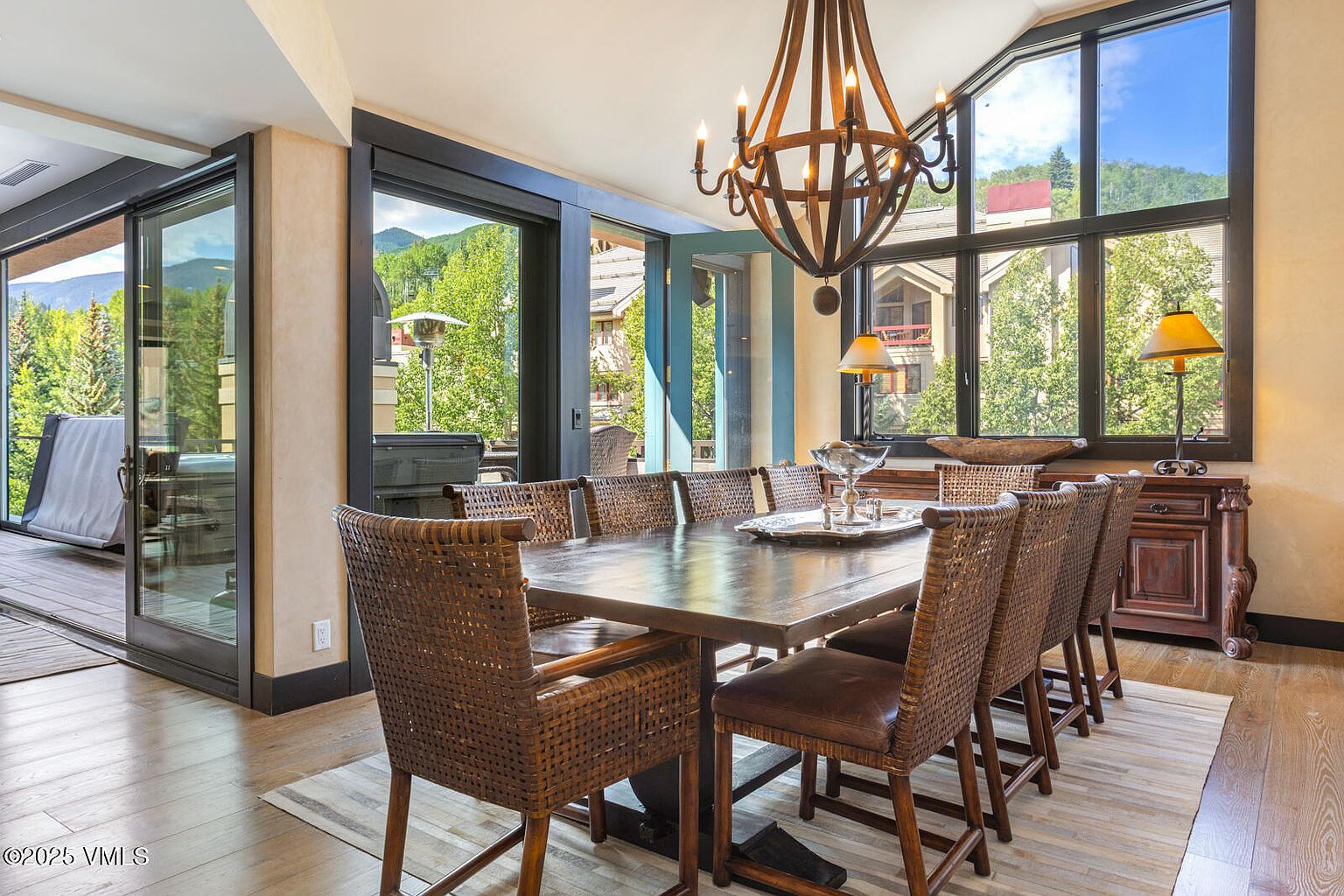 This is an interior shot of a dining room featuring a large wooden table surrounded by woven chairs with leather seats. A rustic chandelier hangs above the table, and a sideboard with lamps sits against the wall. Large windows offer views of the outdoors, and sliding glass doors lead to an outdoor space, creating a bright and inviting atmosphere.