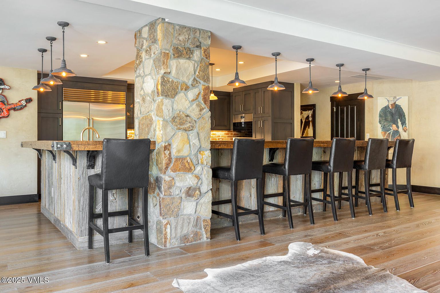 This interior shot showcases a stylish kitchen bar area with a rustic-modern design. The bar features a wooden countertop supported by a stone-clad pillar and reclaimed wood paneling, complemented by black leather bar stools. Overhead, industrial-style pendant lights illuminate the space, adding to the warm and inviting atmosphere.