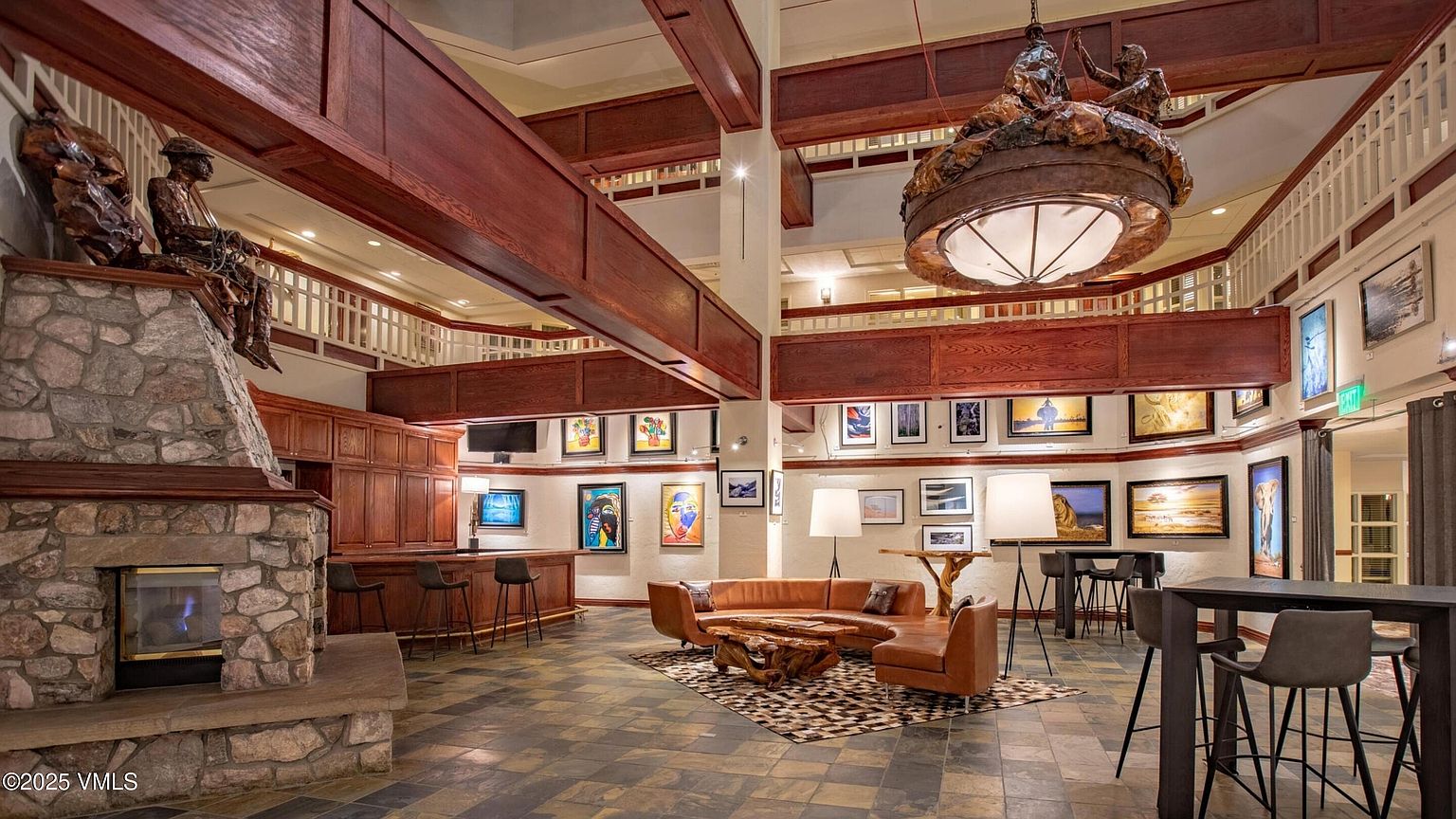 This is an interior shot of a grand living room area, featuring a stone fireplace with a sculpture on top, a seating area with a leather sofa and unique coffee table, and a bar area with stools. The room is decorated with various artworks and illuminated by a large, ornate chandelier, creating a sophisticated and inviting atmosphere.