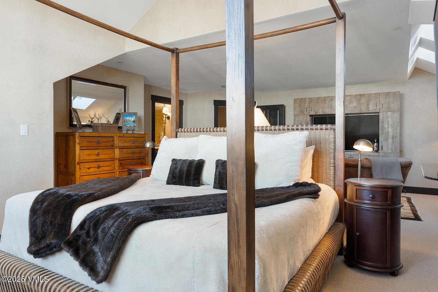 This is a primary bedroom featuring a four-poster bed with a woven headboard and a neutral color palette. The room includes a wooden dresser with a mirror above it, and a round bedside table with a lamp. The overall impression is one of comfort and understated elegance.
