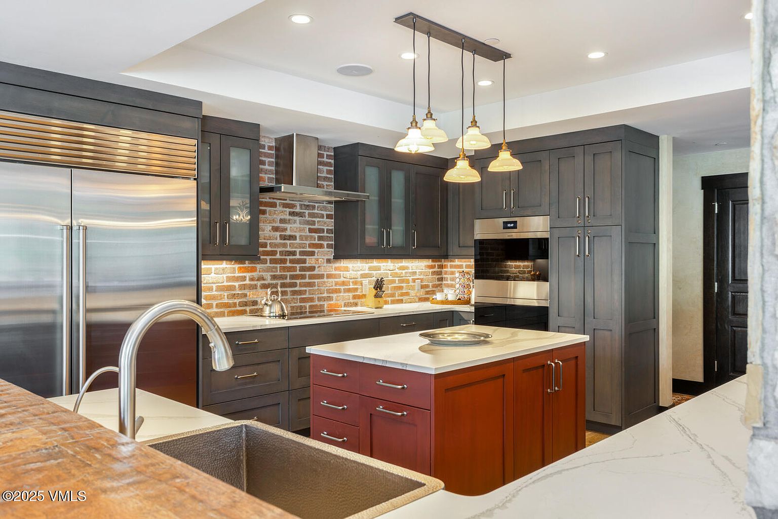 This is a well-appointed kitchen featuring dark gray cabinetry, stainless steel appliances, and a contrasting red island with a white countertop. A brick backsplash adds texture and warmth, while pendant lights provide focused illumination. The kitchen offers a blend of modern and rustic elements, creating a sophisticated and inviting space.