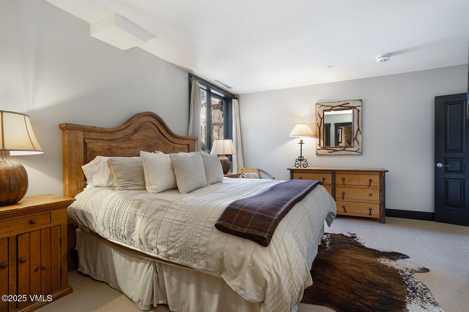 This is a cozy primary bedroom featuring a wooden bed frame with a neutral-toned comforter and decorative pillows. The room includes matching wooden nightstands and a dresser, complemented by lamps that provide soft lighting. A cowhide rug adds a touch of rustic charm to the carpeted floor, and a window with curtains allows natural light to filter in, creating a warm and inviting atmosphere.