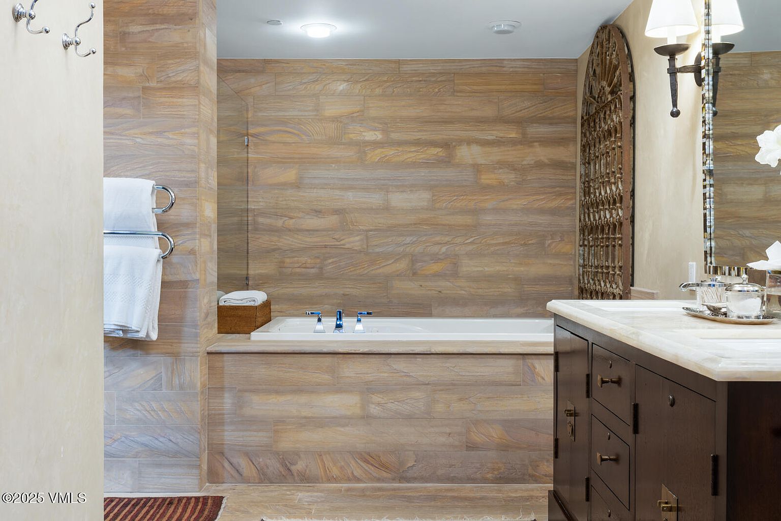 This is a well-appointed primary bathroom featuring a built-in bathtub with a tiled surround that extends to the walls, creating a cohesive and luxurious look. A dark wood vanity with a marble countertop is visible, along with decorative lighting and a large mirror. The overall impression is one of spa-like tranquility and high-end design.